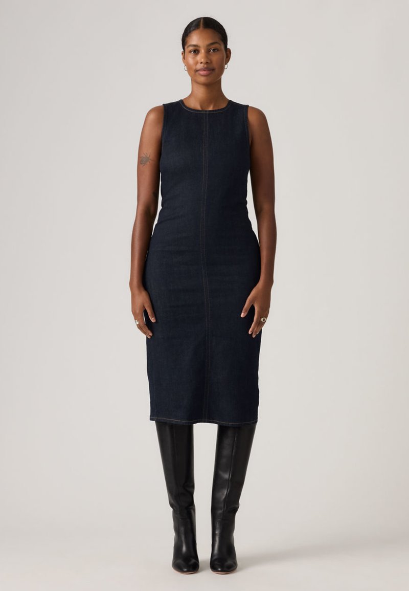 Γυναίκα με σκουρόχρωμα πλεκτά μαλλιά, ντυμένη με αμάνικο μαύρο τζιν φόρεμα μήκους μέχρι τη μέση της γάμπας και μαύρες δερμάτινες μπότες μέχρι το γόνατο, στέκεται μπροστά σε απλό φόντο.