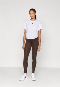 Hvid atletisk top med korte ærmer og sort logo, brune leggings og hvide sneakers. Stoffet er glat og tætsiddende. Enkel, ren design.