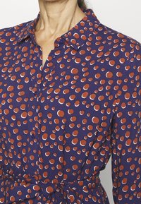 Camicia con bottoni di tessuto viola scuro, caratterizzata da un motivo di grandi pois arancioni e bianchi; include una vita annodata e colletto.