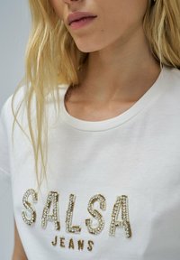 Camiseta de algodón blanca con detalles texturizados y brillantes de "SALSA" y "JEANS" en cristales dorados y transparentes, cuello redondo, mangas cortas.
