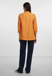 Oranžový blazer s ohrnutými rukávy, s konstrukčně zpracovaným designem. V kombinaci s tmavě modrými, mírně rozšířenými džíny a černými teniskami.