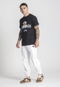 Černé tričko s kreslenou grafikou na přední straně, bílé cargo kalhoty s kapsami a hnědé tenisky s leopardím vzorem a modrými akcenty.