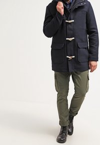 Uomo che indossa un cappotto toggle nero, pantaloni cargo oliva e stivali neri con lacci, mentre cammina con una mano vicino al colletto.