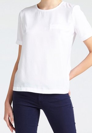 Wit T-shirt met korte mouwen en ronde hals, gedragen met donkerblauwe broek, handen rustend op de heupen tegen een effen lichte achtergrond.
