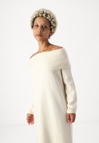 Λευκό-κρέμ μάλλινο φόρεμα με ραβδωτό ντεκολτέ που αφήνει τους ώμους ακάλυπτους και μακριά μανίκια. Το ύφασμα έχει απαλή, υφασμένη επιφάνεια.