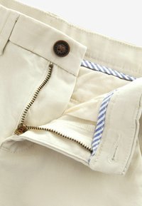 Pantalones chinos de algodón beige con cierre de metal y botón. El interior cuenta con un forro a rayas en azul y blanco y detalles de costura en contraste.