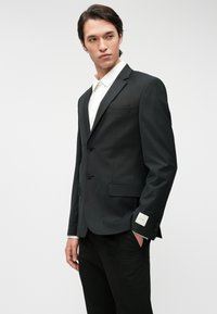 Blazer preto com dois botões, um único bolso no peito e camisa de dress branco em contraste. Calças pretas com uma textura suave.