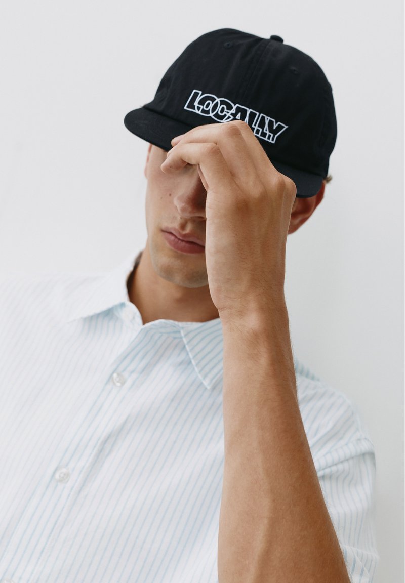 Jeune homme portant une casquette noire avec le texte "LOCALLY" et une chemise blanche à boutons, couvrant légèrement son visage avec la main.