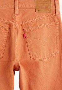 Jean en denim orange avec une étiquette en cuir, deux poches arrière ornées d'arcs cousus, et une petite étiquette rouge sur la poche droite.