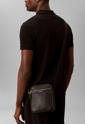 Homme portant une chemise noire à manches courtes, avec un sac bandoulière noir Calvin Klein muni d'une poche avant zippée, porté sur l'épaule droite.