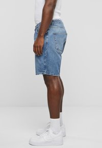 Shorts en denim de lavage bleu clair, arrivant au genou, avec une coupe classique, cinq poches et un ourlet droit, portés avec des baskets blanches et des chaussettes.
