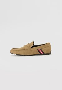 Mocasin de ante marrón con suela plana, que presenta una correa trenzada y un acento lateral en rayas rojas y negras. Textura suave y diseño sencillo.