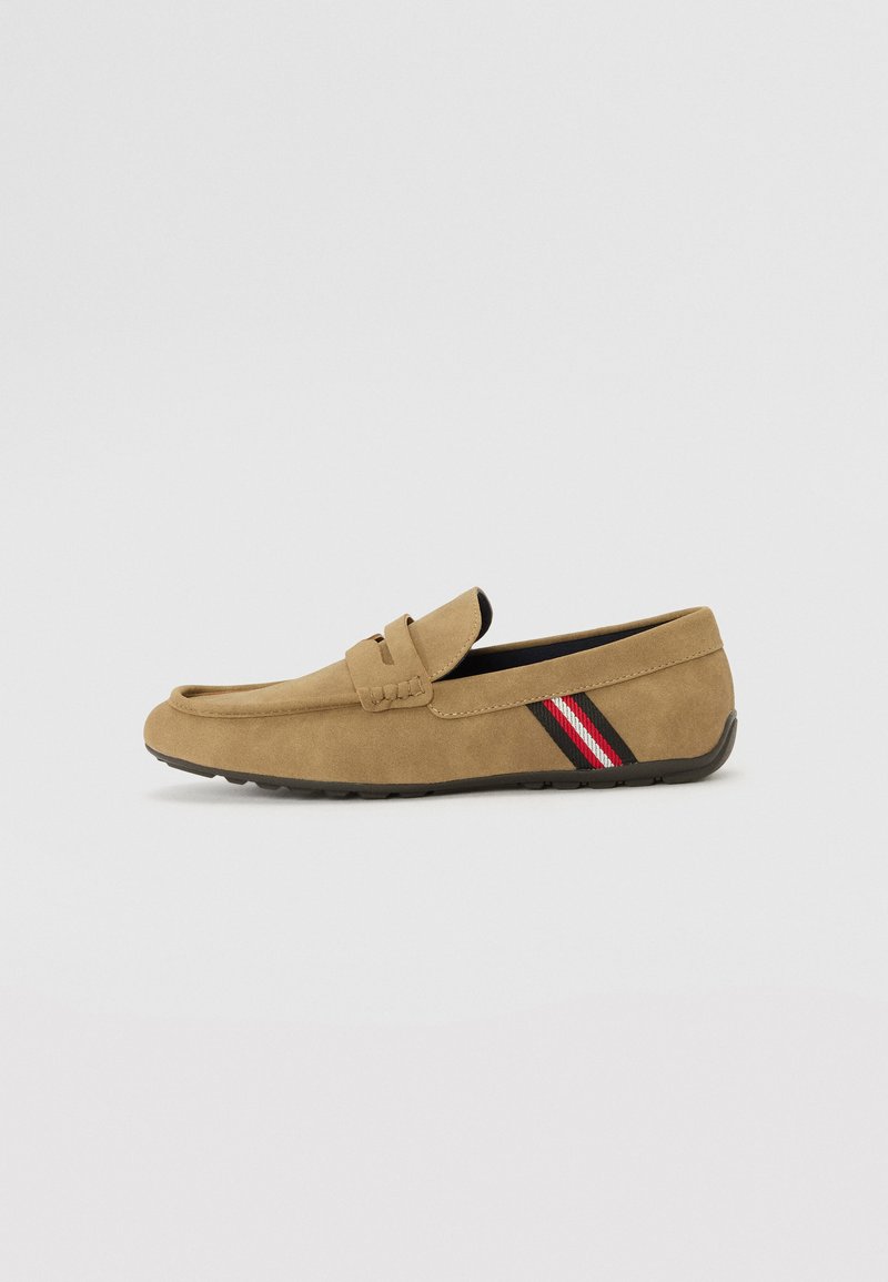 Mocasin de ante marrón con suela plana, que presenta una correa trenzada y un acento lateral en rayas rojas y negras. Textura suave y diseño sencillo.