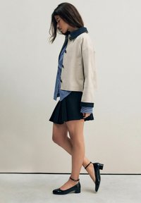 Beige kurz geschnittene Jacke mit einem dunkelblauen Kragen, kombiniert mit einem blauen gestreiften Hemd und einem schwarzen Faltenrock. Schwarze Lackleder-Absatzschuhe.