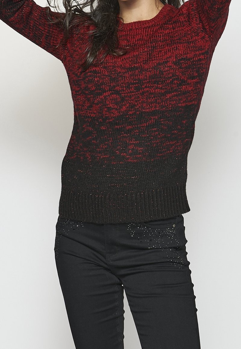 Personne portant un pull en maille dégradé rouge et noir et un pantalon noir avec de petites décorations brillantes, bras levés au-dessus de la tête.