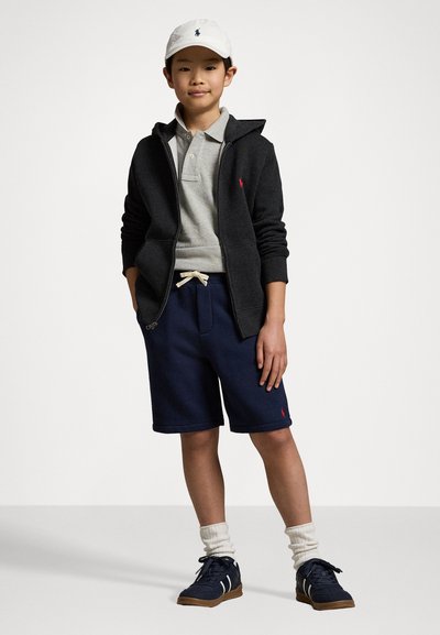 Γκρι πόλο μπλούζα κάτω από μαύρο φούτερ με φερμουάρ, ναυτικά σορτς, άσπρο καπέλο και ναυτικά αθλητικά παπούτσια με καφέ σόλες. Ριγέ άσπρες κάλτσες. Κόκκινες λεπτομέρειες λογότυπου.