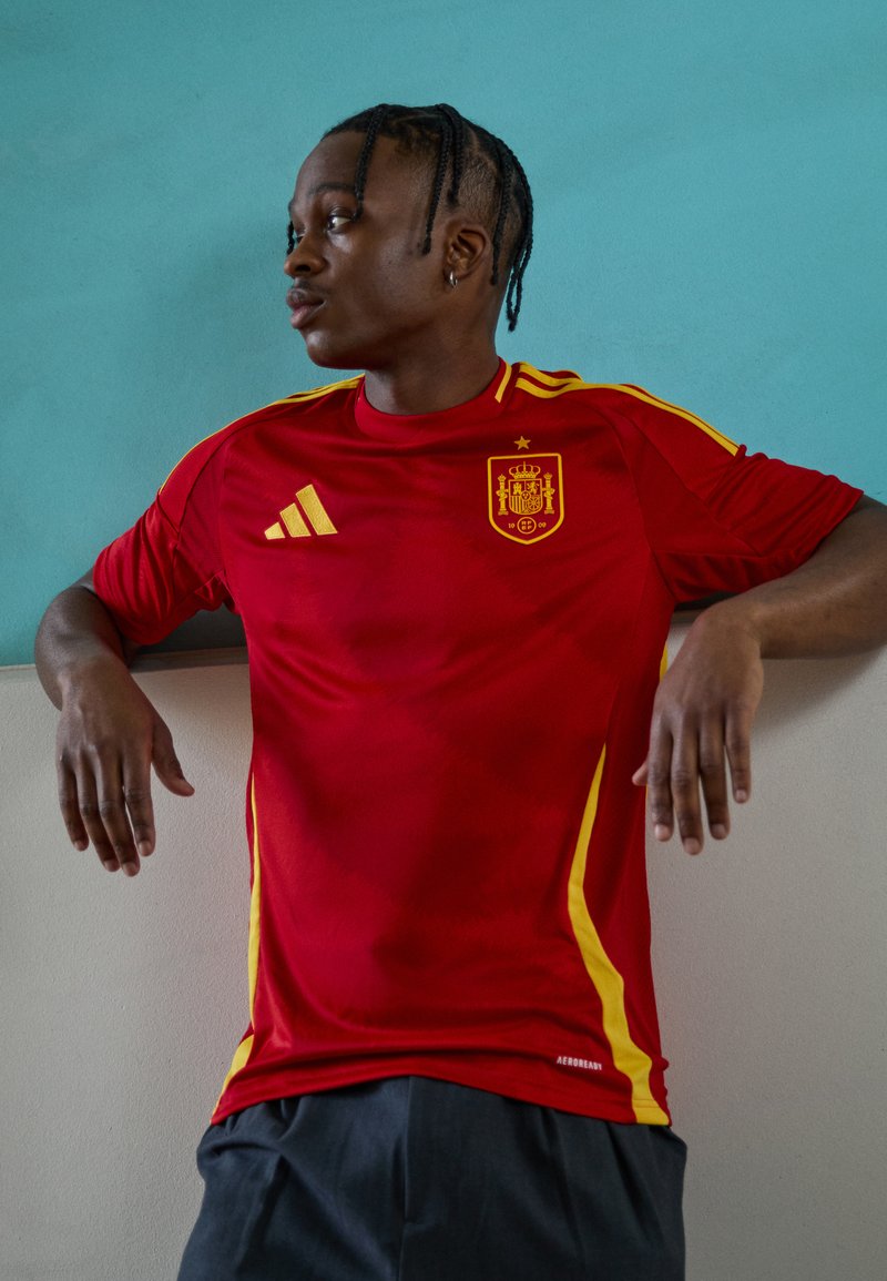Crvena atletska majica s kratkim rukavima i žutim detaljima, okrugli izrez. Sadrži španjolski grb i Adidasov logo. Glatka tekstura tkanine.