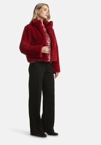 Giacca in ecopelliccia rossa con collo alto, vestibilità ampia e maniche larghe, abbinata a una maglia stampata e pantaloni neri a gamba larga.