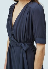 Tyrkysově modré zavinovací šaty s krátkým rukávem, s plisovanými detaily na ramenou a uvázaným páskem v pase. Hladká, lehká tkanina.