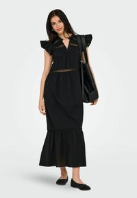 Black maxi dress with ruffled sleeves, keyhole neck, lace accents at waist, and a flared hem. Model holding a black tote bag and wearing flats.
