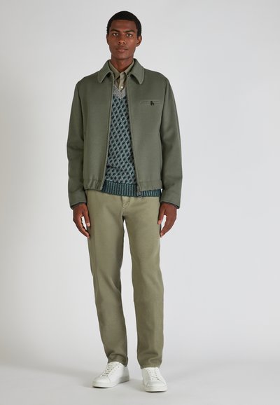Grüne Wollmischjacke mit Kragen, getragen über einem gemusterten blau-grünen Pullover, kombiniert mit hellgrünen Hosen und weißen Sneakers.