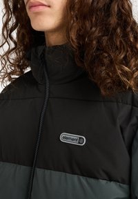 Persona con capelli ricci che indossa un giubbotto impermeabile nero e verde scuro con colletto alto e un logo "element" sul petto.