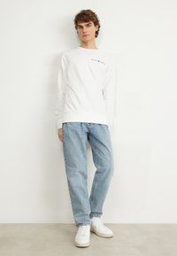 Hvid langærmet t-shirt med "GANT 1949" logo på brystet, parret med lyseblå jeans og hvide sneakers. Glat stof, afslappet pasform.