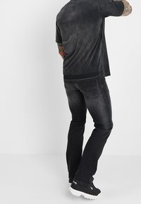 Uomo in piedi con la schiena rivolta verso la camera, indossa jeans neri scoloriti, una t-shirt grigia scura e sneakers nere FILA con suole bianche.