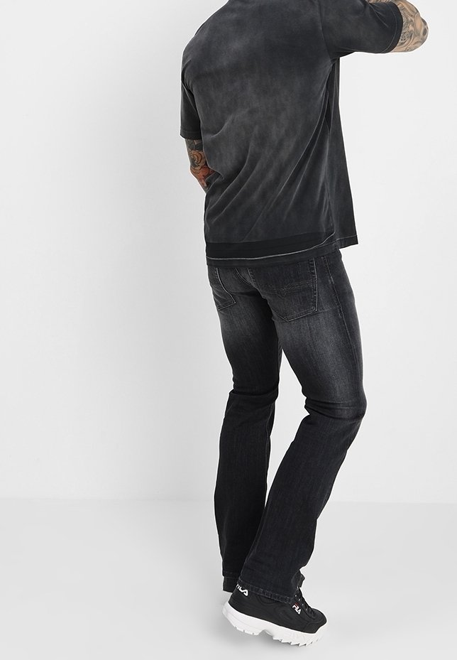 Uomo in piedi con la schiena rivolta verso la camera, indossa jeans neri scoloriti, una t-shirt grigia scura e sneakers nere FILA con suole bianche.