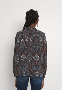 Chemise à manches longues de couleur bleu foncé, ornée d'un motif cachemire avec des accents blancs, orange et bleu canard. Détail en forme de trou de serrure à l'encolure arrière.