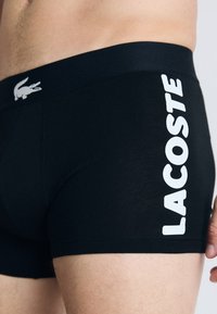 Zwarte boxershorts van rekbare stof met een strakke pasvorm. Heeft witte "LACOSTE" branding op de zijkant en een klein krokodillogo op de tailleband.