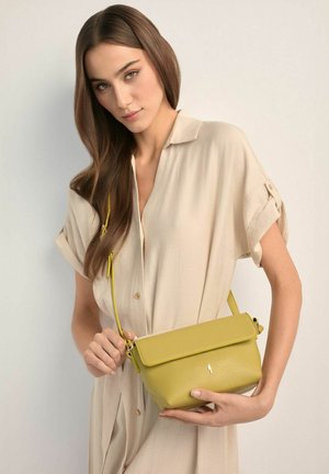 Kobieta w beżowej sukience zapinanej na guziki, trzymająca małą żółtą skórzaną torebkę na ramię z klapką i paskiem na ramię na jednolitym tle.