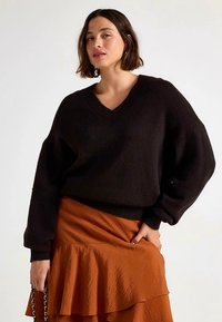 Schwarzer Strickpullover mit V-Ausschnitt, lockerem Sitz und voluminösen Ärmeln, kombiniert mit einem gestuften, rostfarbenen Rüschenrock.