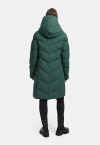 Grön pufferjacka med en stor huva, med chevronstygn, knälång design, och en del av blanka svarta stövlar syns i botten.