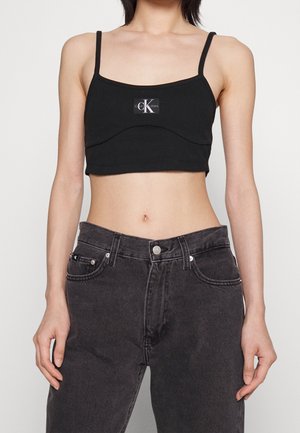 Osoba ubrana w czarny krótki top Calvin Klein z cienkimi ramiączkami oraz ciemnoszare dżinsy z wysokim stanem, stojąca na jednolitym tle.