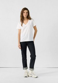 T-shirt bianco a scollo a V, jeans in denim scuro con risvolti, e sneakers argentate. Il modello si trova in piedi con le mani rilassate ai lati su uno sfondo semplice.