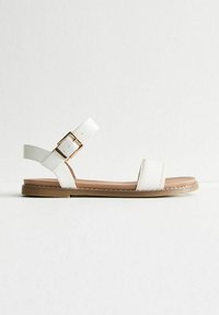 Sandales plates blanches avec une semelle marron, présentant une large sangle à l'avant et une sangle de cheville réglable avec une boucle couleur or.