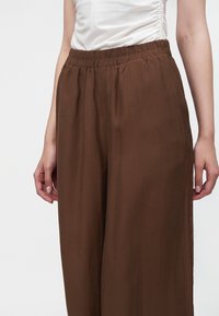 Femme portant un pantalon marron ample taille haute avec une ceinture élastique et un haut blanc froncé sans manches sur un fond uni.