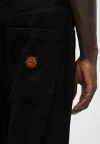 Melni denima bikses ar izteiksmīgu sarkanu un dzeltenu "Santa Cruz" logo uz aizmugurējā kabatas. Gluda virsma, klasiskā piegriezuma.