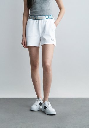 Femme portant un short blanc EA7 avec une ceinture élastique et des baskets blanches avec des accents noirs, debout devant un fond gris uni.