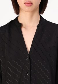 Black blouse from a lightweight fabric featuring a dotted pattern, placket with buttons, and a scalloped collar detail.