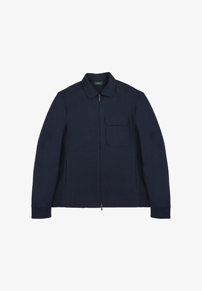 Granatowa kurtka zapinana na zamek, wykonana z teksturowanej tkaniny, z klasycznym kołnierzem oraz kieszenią na piersi z kwadratową klapą. Prostokątna sylwetka.