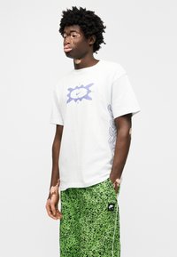 Fehér póló lila Nike logóval és grafikával. Zöld mintás rövidnadrággal párosítva, melyen fekete Nike címke található.