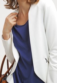Blazer bianco con design moderno e aperto; indossato sopra una maglia blu navy. Dettaglio zip notevole sulla cucitura laterale del blazer. Borsa in pelle visibile.