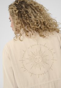 Chaqueta beige con un sol y texto bordados en la parte posterior. Presenta acentos cosidos y una tela suave y texturizada.