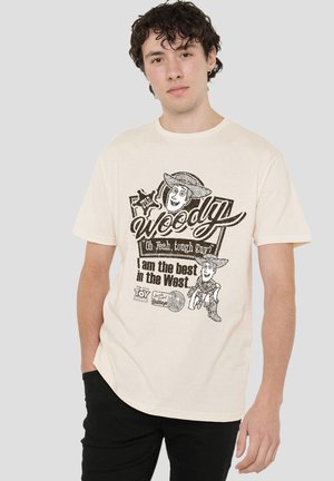 Young man wearing a beige Toy Story Woody graphic t-shirt with text "Oh yeah, tough guy? I am the best in the West."