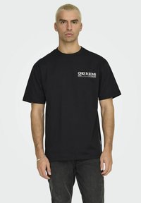 T-shirt in cotone nero con collo a giro, maniche corte e un logo bianco stampato sul lato sinistro del petto. Design semplice, vestibilità comoda.