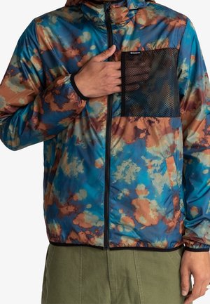 Person trägt eine blaue und braune Batikjacke mit schwarzer Netz-Brusttasche, grüne Hose und hat die Hand in der Tasche.