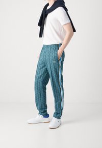 Joggingové kalhoty zelenomodré barvy s bílými a modrými pruhy na bocích, elastickým pasem a jemným logem, doplněné bílým tričkem.