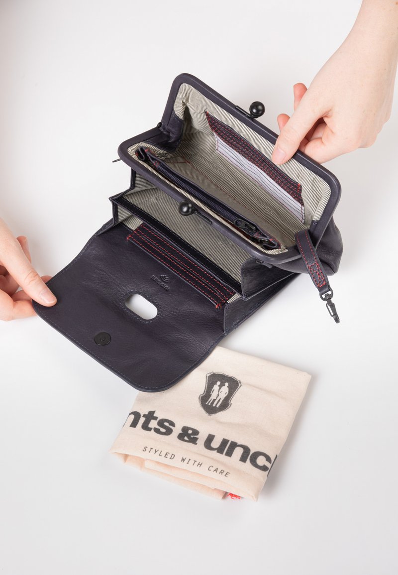 Portafoglio in pelle blu navy con scomparti interni e una fodera a strisce. Una borsa di stoffa con logo è visibile sotto il portafoglio. Chiusura con bottone automatico.